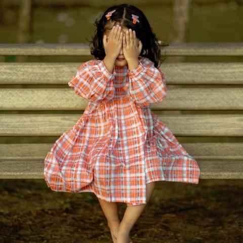 A young barefoot girl in a plaid orange dress sits on a bench, covering her eyes with her hands, playing hide-and-seek.