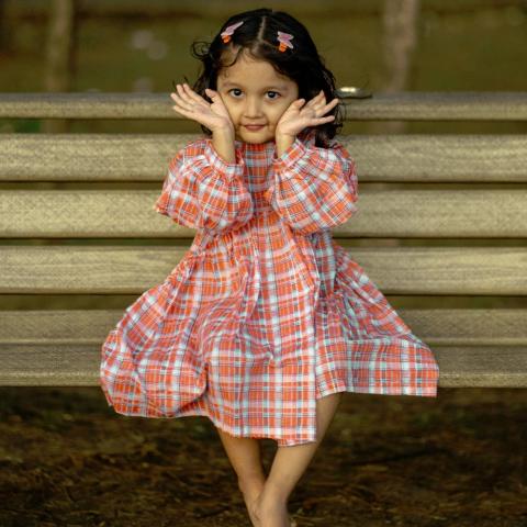 A young girl in a plaid orange dress sits barefoot on a bench, hands cupping her face in a playful pose.