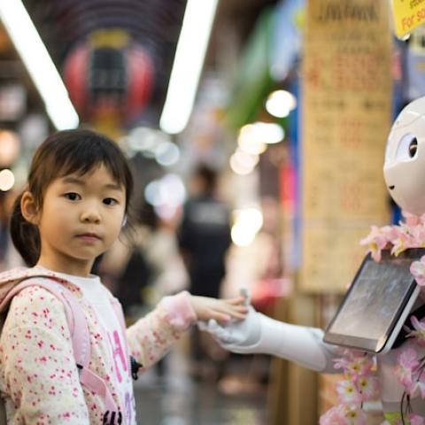A young girl with a backpack touches the hand of a white, humanoid robot in a busy market.